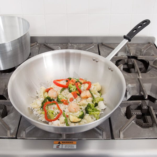 A Vollrath stainless steel stir fry pan on a stove with food in it.