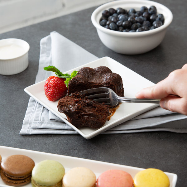 A hand holding a fork to a white square porcelain plate with chocolate cake.