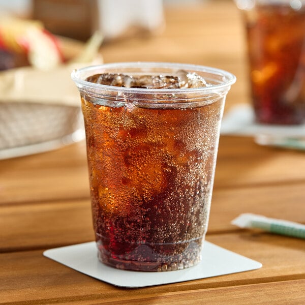 A clear 12 oz plastic cold cup filled with a dark carbonated beverage and ice, placed on a coaster.