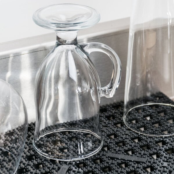 A clear glass jug with a handle on a San Jamar black interlocking bar mat with glass cups.