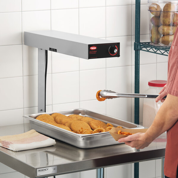 A stainless steel overhead food warmer mounted above a tray of fried foods in a commercial kitchen setting.