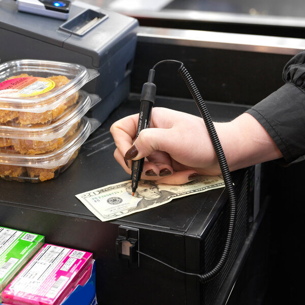 A person is using a counterfeit detection marker to check a $20 bill at a checkout counter, with packaged food items and a cash register visible nearby.