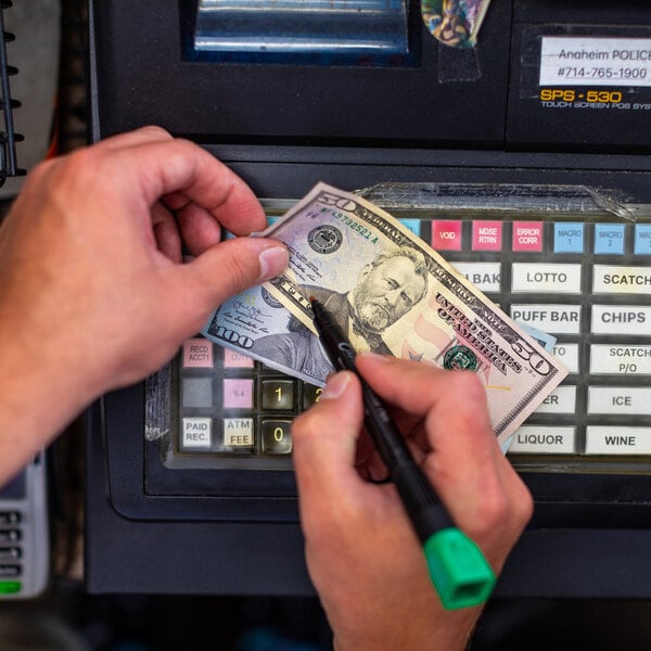 A person is using a marker to check a $50 bill in front of a cash register with labeled buttons.