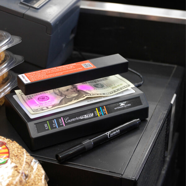 A black Dri Mark Tri-Test UV Counterfeit Detection System with a counterfeit detection pen and U.S. currency placed under the UV light.