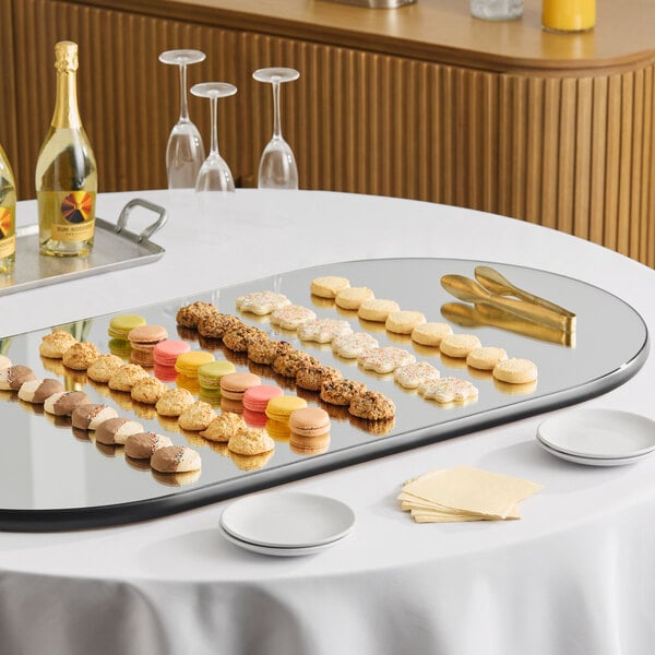 An oval rimless mirror food display tray measuring 24 by 48 inches, shown with assorted desserts arranged on top.