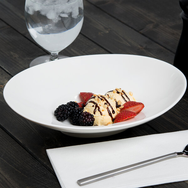 A white 10 Strawberry Street Whittier shallow oval porcelain bowl on a table with a glass of water and a long silver spoon.