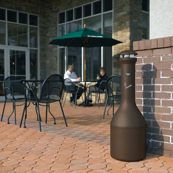 A Rubbermaid brown cylindrical free standing cigarette receptacle on an outdoor patio.