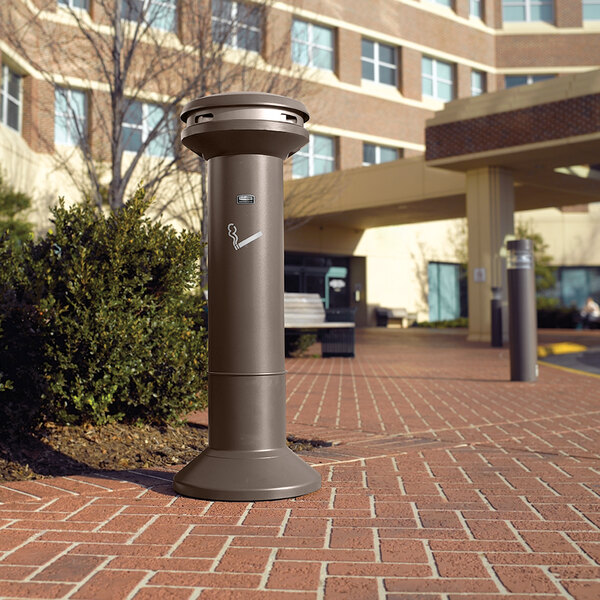 A Rubbermaid Infinity Aged Bronze cigarette receptacle on a brick walkway.
