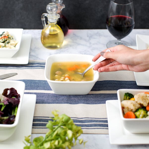 A person holding a 10 Strawberry Street Whittier white square bowl with a spoon over soup.