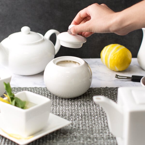 A person pouring sugar into a white teapot.