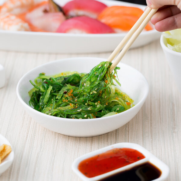 A pair of chopsticks holding seaweed salad over a white 10 Strawberry Street sauce dish.