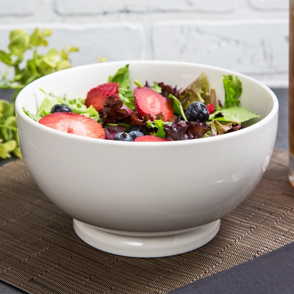 A white round porcelain footed bowl filled with a salad with strawberries and blueberries.