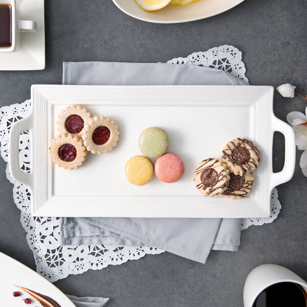 A white rectangular porcelain platter with black circles on the handles holding a plate of cookies and tea.