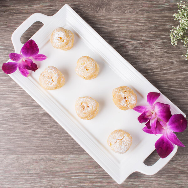 A white 10 Strawberry Street rectangular porcelain tray with handles on a table with donuts and flowers.