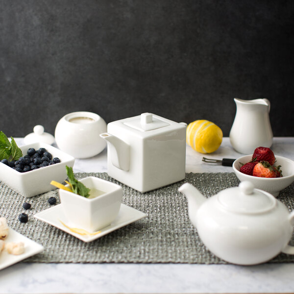 A white square teapot with a lid on a table set with white dishes and fruit.
