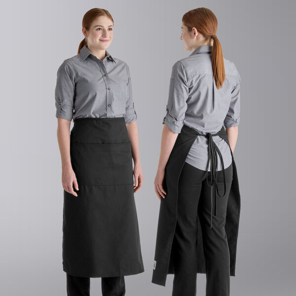 Two people are shown wearing long black aprons with two front pockets over gray button-up shirts and black pants, with one person facing forward and the other showing the back view.