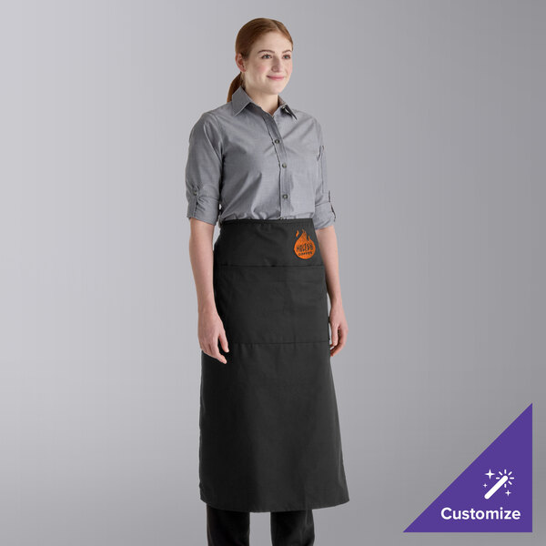 A person is wearing a long black apron with two pockets over a gray button-up shirt, standing against a plain background.