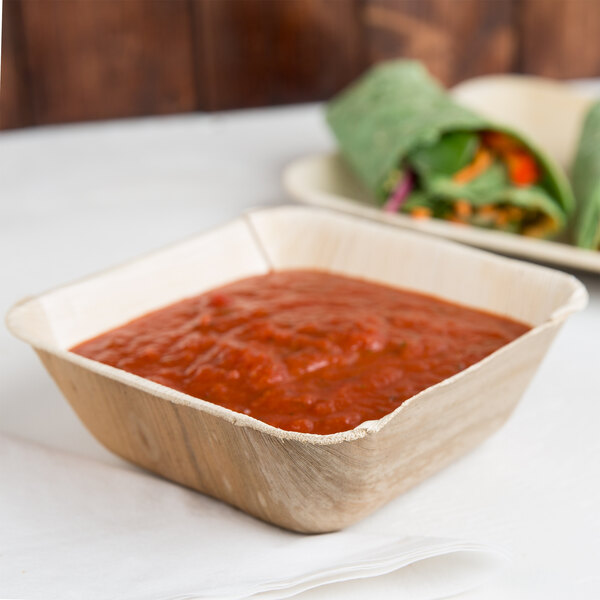 A white table with Eco-gecko palm leaf bowls of tomato sauce and burritos.