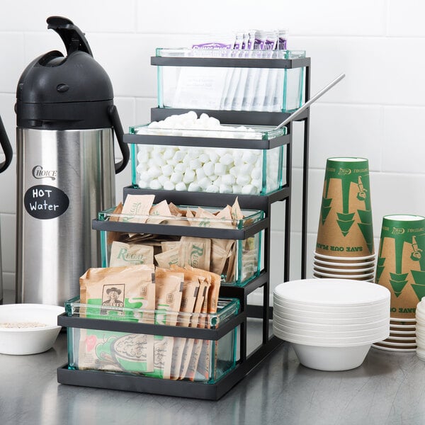 A black Cal-Mil stair step condiment display with glass jars on a counter with a coffee maker and coffee containers.