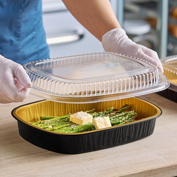 A person holding a Durable Packaging foil entree pan with a plastic lid over asparagus and cheese.