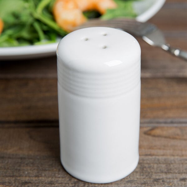 A close-up of a Tuxton white china pepper shaker on a table.