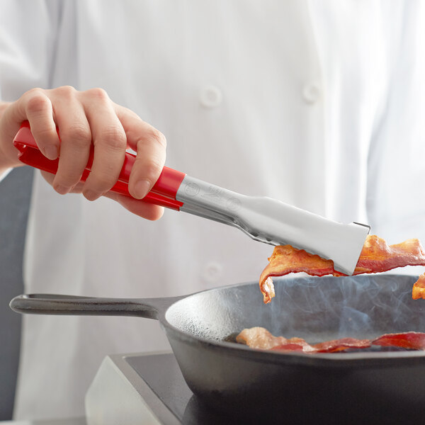 A hand using Vollrath VersaGrip tongs to cook bacon in a pan.