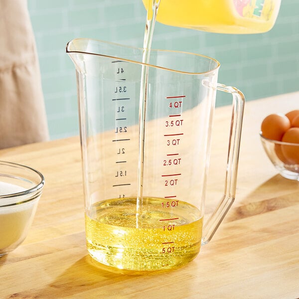 A person pouring yellow liquid into a Cambro clear polycarbonate measuring cup on a counter in a home kitchen.