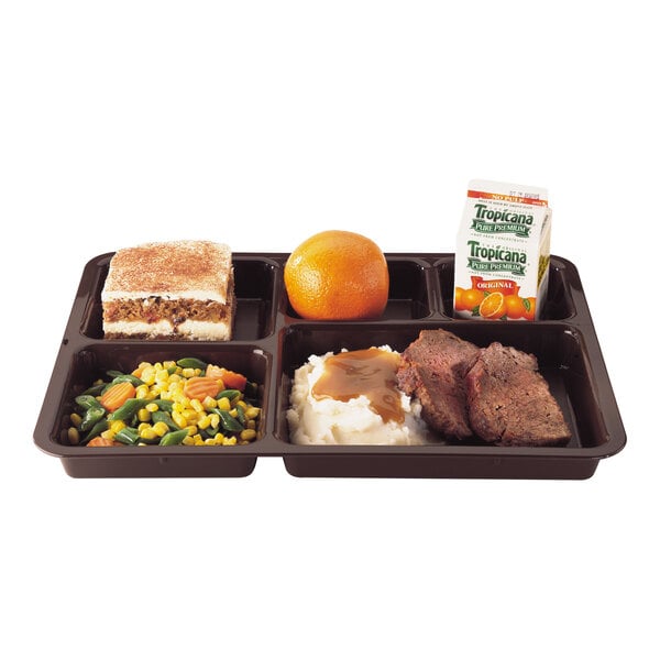A compartmentalized brown tray containing a slice of cake, a whole orange, a carton of orange juice, mixed vegetables, mashed potatoes with gravy, and two pieces of meat.