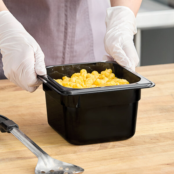 A person in gloves holding a black Cambro H-Pan container of macaroni and cheese.