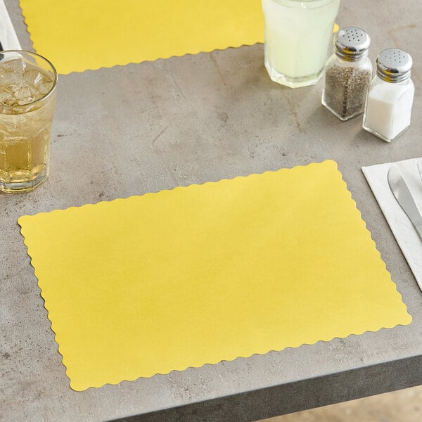 A yellow scalloped paper placemat on a table with a glass of liquid.