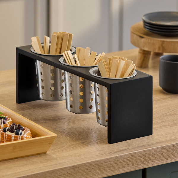 A three-cylinder horizontal flatware or condiment display with a black frame and perforated metal containers holding wooden utensils.
