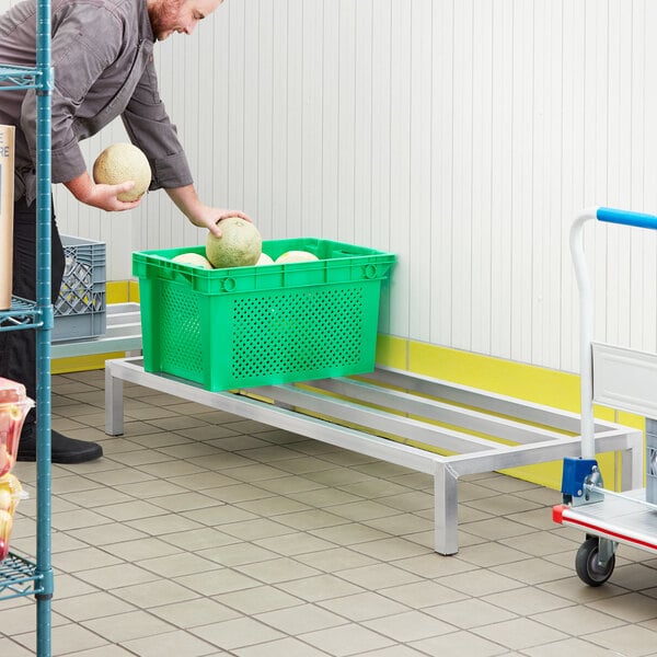 A man pushing a cart with a Regency aluminum dunnage rack holding a green basket of melons on a metal platform.