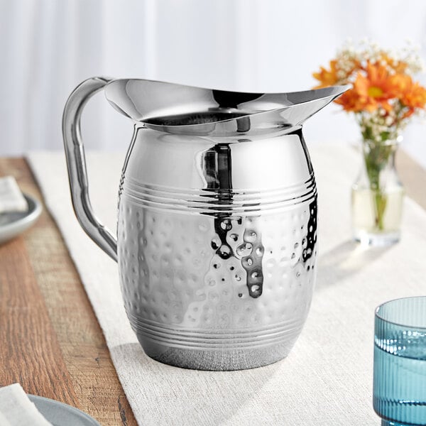 An American Metalcraft hammered stainless steel water pitcher on a table with a white tablecloth.