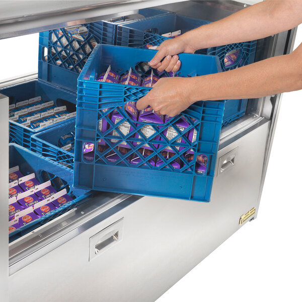 A person using a blue Traulsen milk crate to fill a blue Traulsen milk cooler on a counter in a school kitchen.