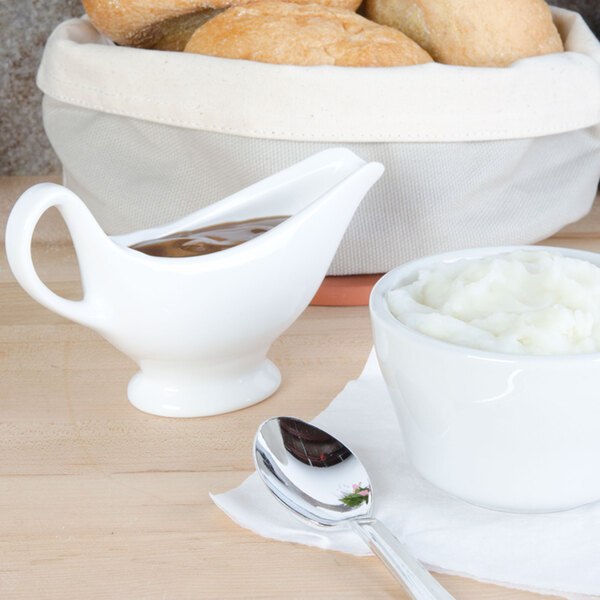 A white American Metalcraft gravy boat with brown gravy next to a bowl of mashed potatoes with a spoon.