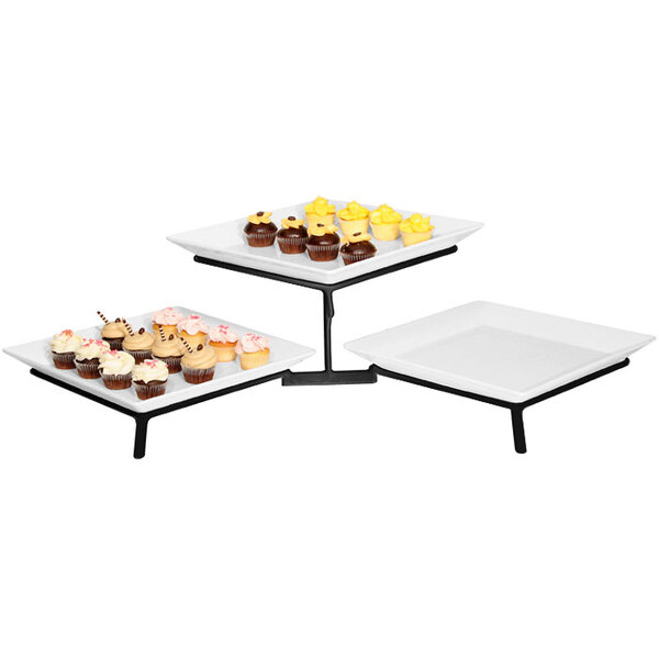 A black Cal-Mil two tier wire stand with white square trays holding cupcakes on a table.