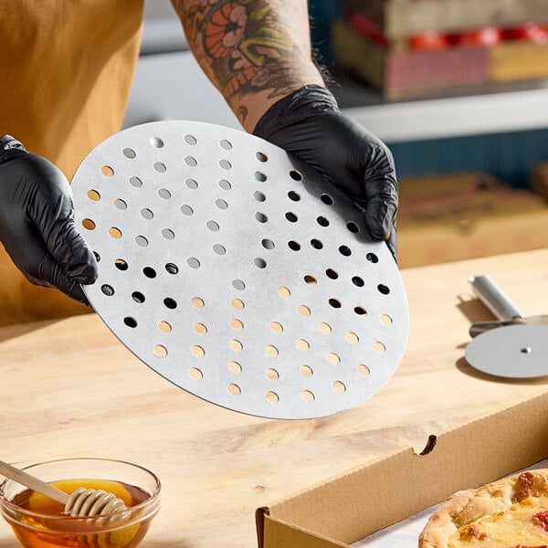 A round, perforated metal pizza disk being held above a wooden surface.