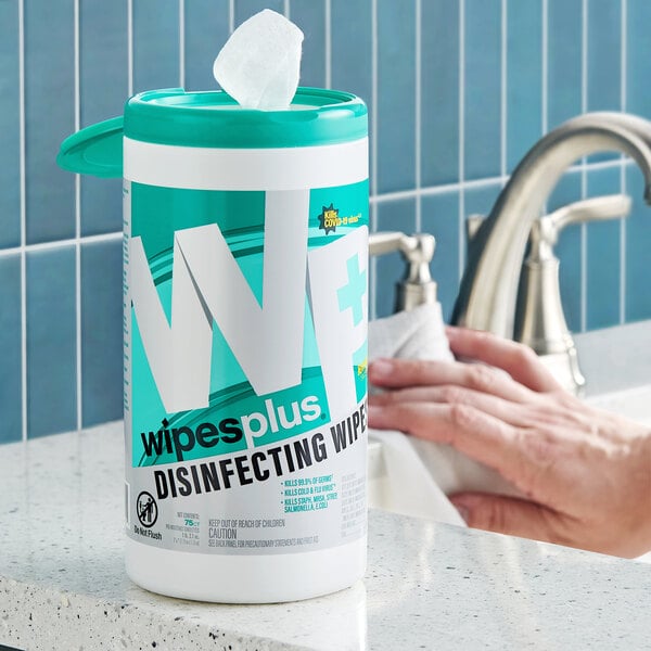 A container of disinfecting wipes is placed on a bathroom counter next to a sink, with a person using a wipe to clean the surface.