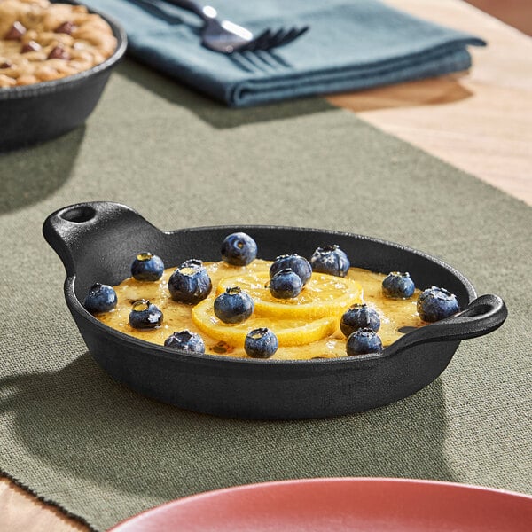 A small, round, black cast iron casserole dish with handles on both sides, filled with a dessert topped with blueberries and lemon slices.