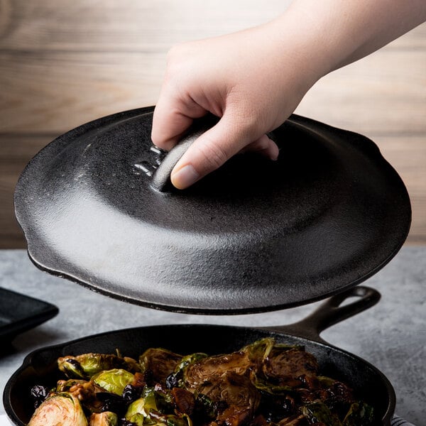 A hand holding a Lodge cast iron lid over a pan of brussels sprouts.