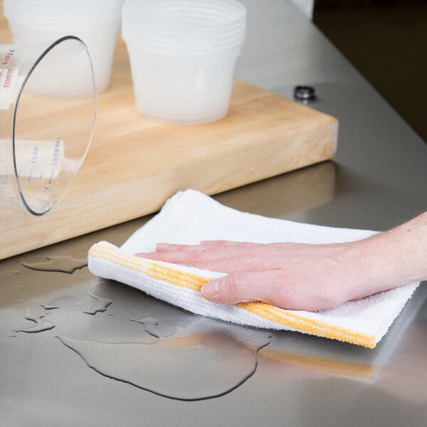 A hand holding a white Chef Revival bar towel cleaning a glass on a counter.