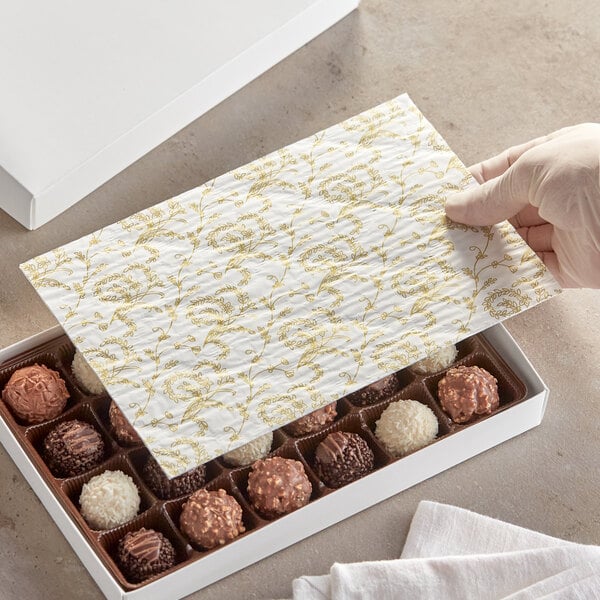 A person's hand holding a white glassine paper pad with a gold floral pattern over a box of chocolates.