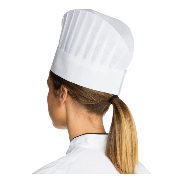 A person wearing a tall, white pleated chef hat and a white chef coat, viewed from behind.