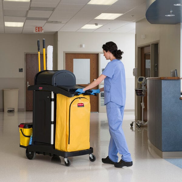 A nurse in blue scrubs pushing a Rubbermaid janitor cart with a locking hood and cabinets.