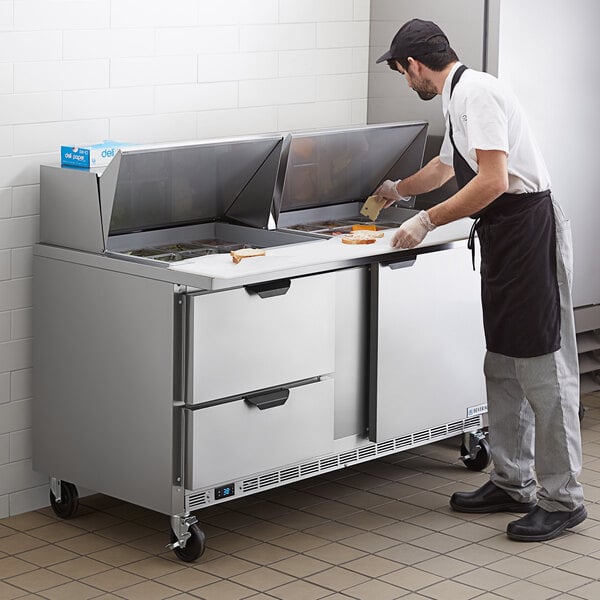 A man in a black apron and hat and gloves cutting cheese on a Beverage-Air sandwich prep table.