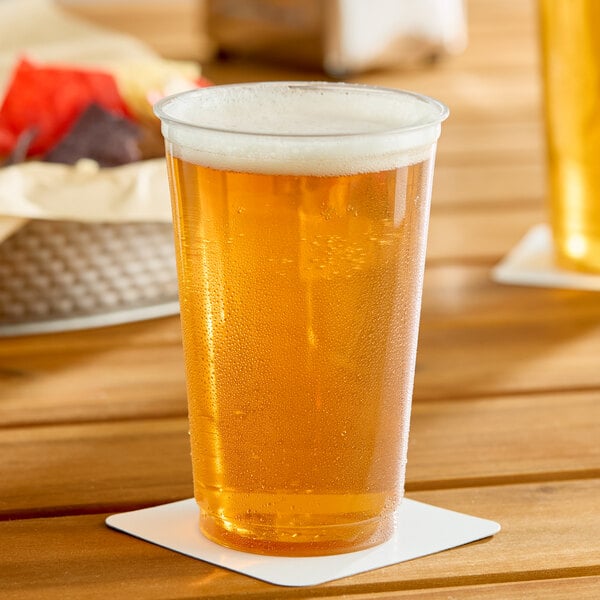 A clear 20 oz. plastic cold cup filled with a light amber beverage and topped with foam, placed on a white coaster.