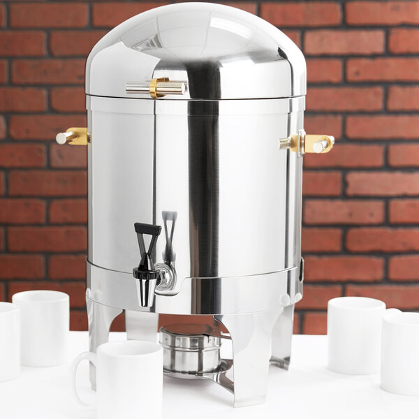 A Vollrath stainless steel coffee urn with brass trim on a table with white cups.