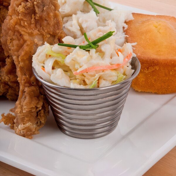 A plate with fried chicken, cornbread, and a stainless steel round ribbed sauce cup filled with coleslaw on a table.