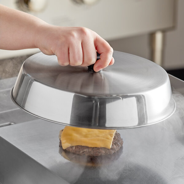 A hand holding an American Metalcraft stainless steel basting cover over a cheeseburger on a grill.
