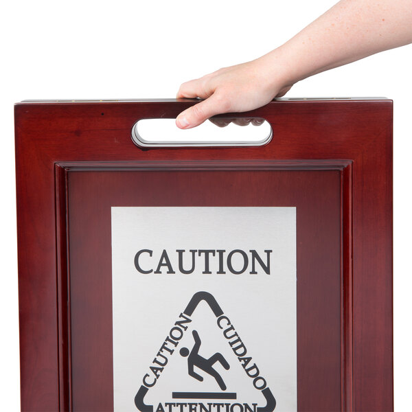 A hand holding a Rubbermaid wooden wet floor sign that says "Caution"